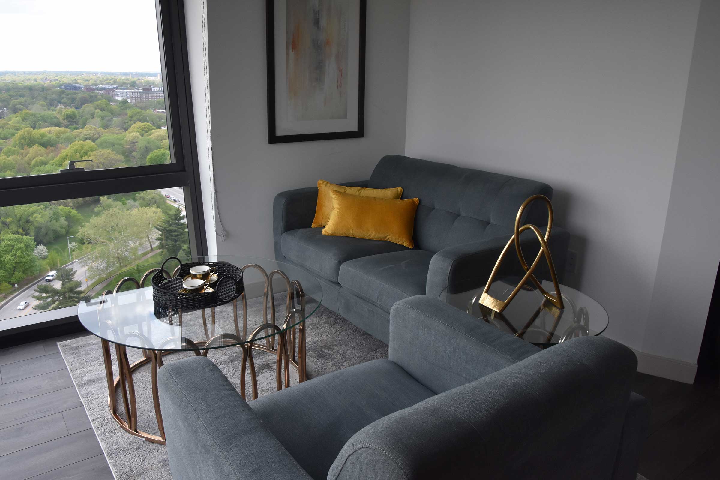 two grey modern style couches sit caddy corner to one another in the living room of an apartment with floor to ceiling windows. There is a glass coffee table that sits between them with chic modern accent items displayed on top. 