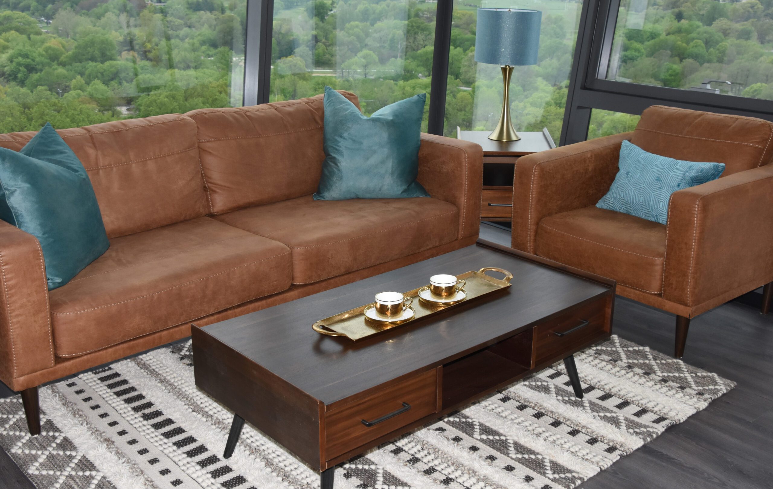 a brown leather couch and chair sit caddy corner to one another with a stylish wood coffee table between them. the couches both have bright turquoise pillows in the corners. the setting is a high-rise apartment building.